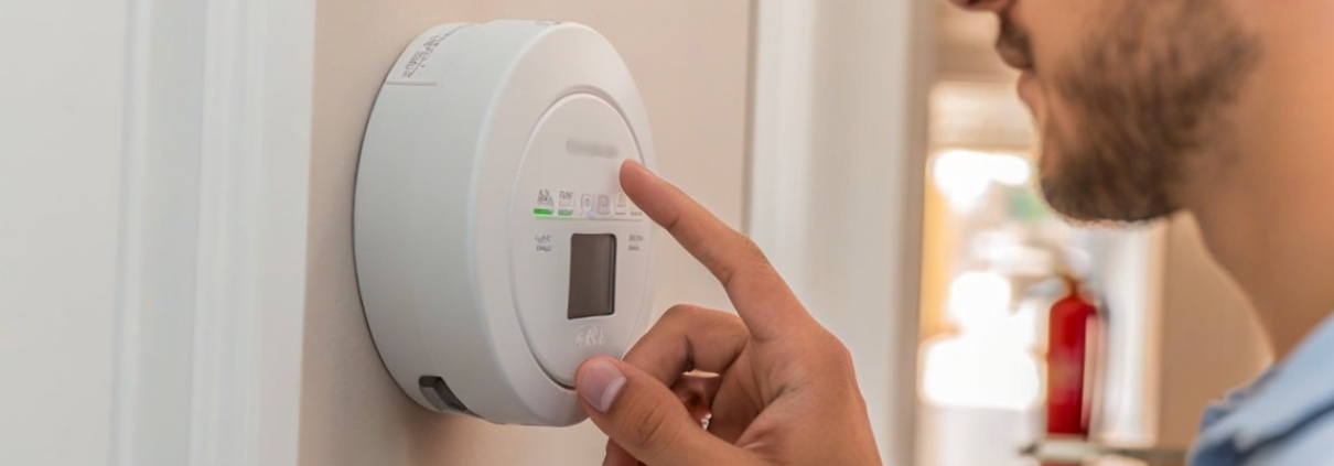 A close-up of a person testing a white, round-shaped carbon monoxide detector mounted on a hallway wall. The individual is pressing the test button on the detector, showing a look of concentration. The background subtly features other safety elements like a faraway fire extinguisher and a smoke detector, reinforcing the theme of comprehensive home safety measures. The lighting is bright and clear, giving the scene a practical and informative feel. no fire in the background.