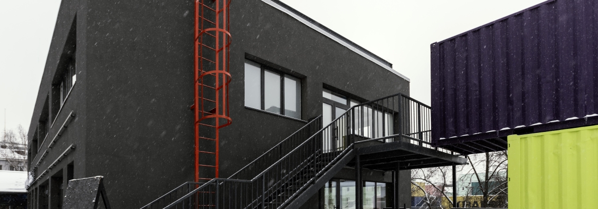 A modern building with a prominent red escape ladder attached to the exterior wall, leading to the roof. The building also features a black metal staircase, creating a secondary emergency exit route. The scene is set in winter, with snow covering the ground, emphasizing the importance of accessible escape routes in all weather conditions.