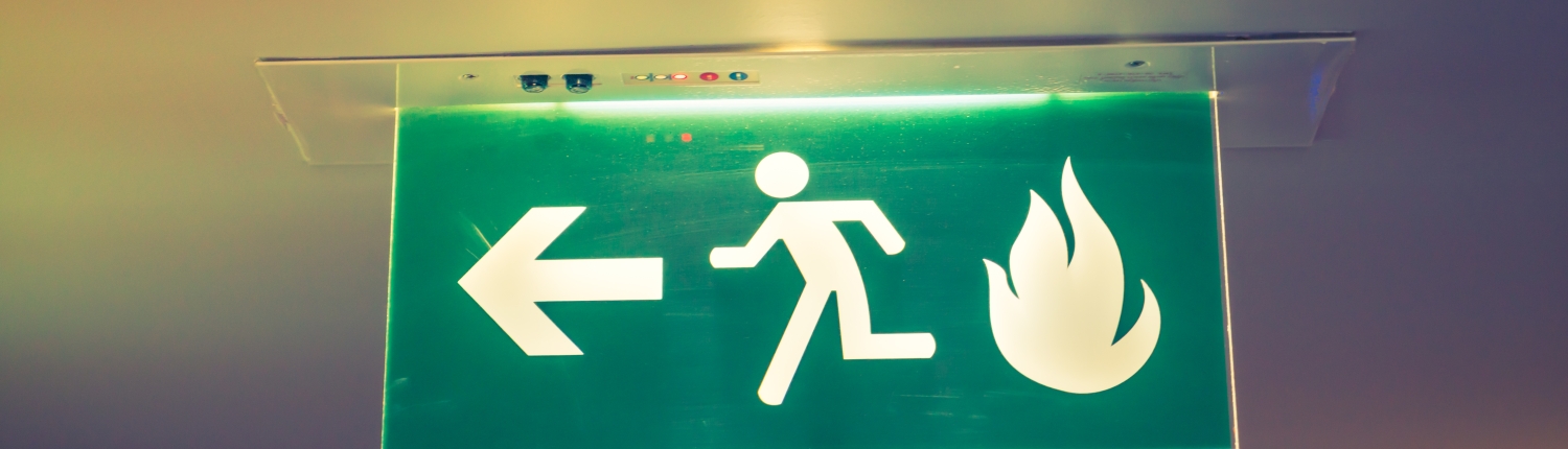 A green emergency exit sign mounted on the ceiling, featuring a white icon of a running person and an arrow pointing left towards the exit. A flame symbol is also present, indicating the direction for safe evacuation in case of a fire. The sign is illuminated, enhancing its visibility in low-light conditions.